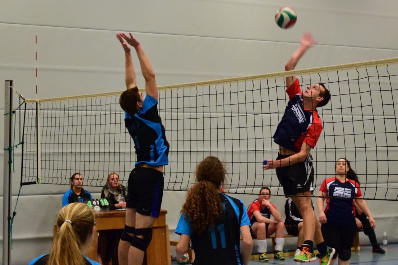 Spielszene im Volleyball mit einem Spieler, der den Ball über das Netz anfeuert, während ein Gegner blockt.