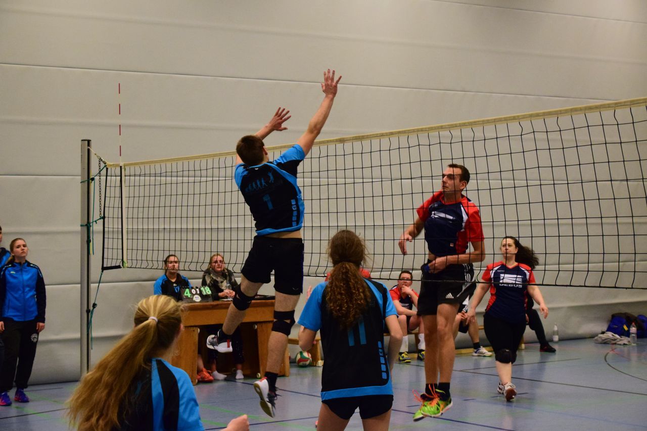 Spielszene eines Volleyballmatches mit Spielern in blauer und roter Sportbekleidung, Netzspannung sichtbar.