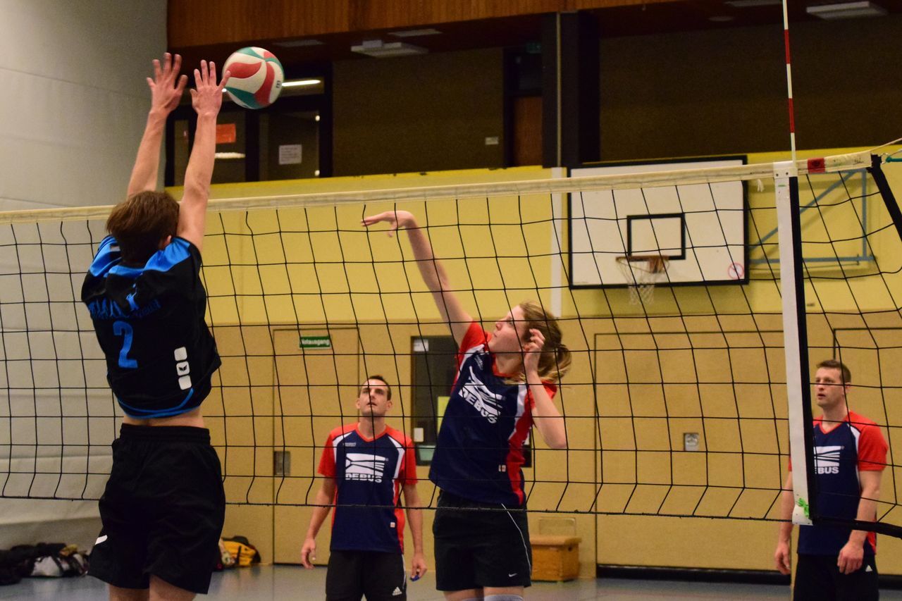Spieler springen zum Blocken eines Volleyballs über das Netz in einer Sporthalle während eines Spiels.