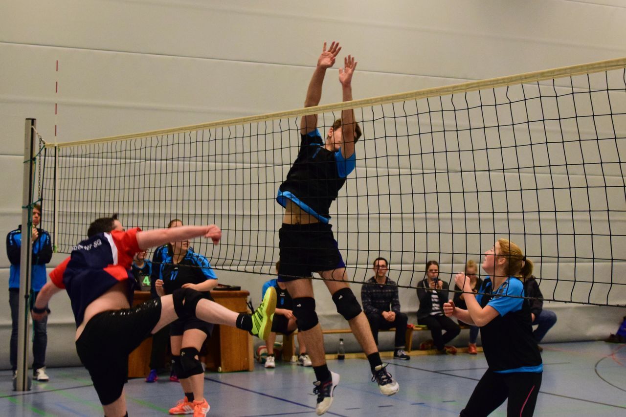 Ein Volleyballspieler springt, um einen Ball über das Netz zu blocken, während Zuschauer im Hintergrund zuschauen.