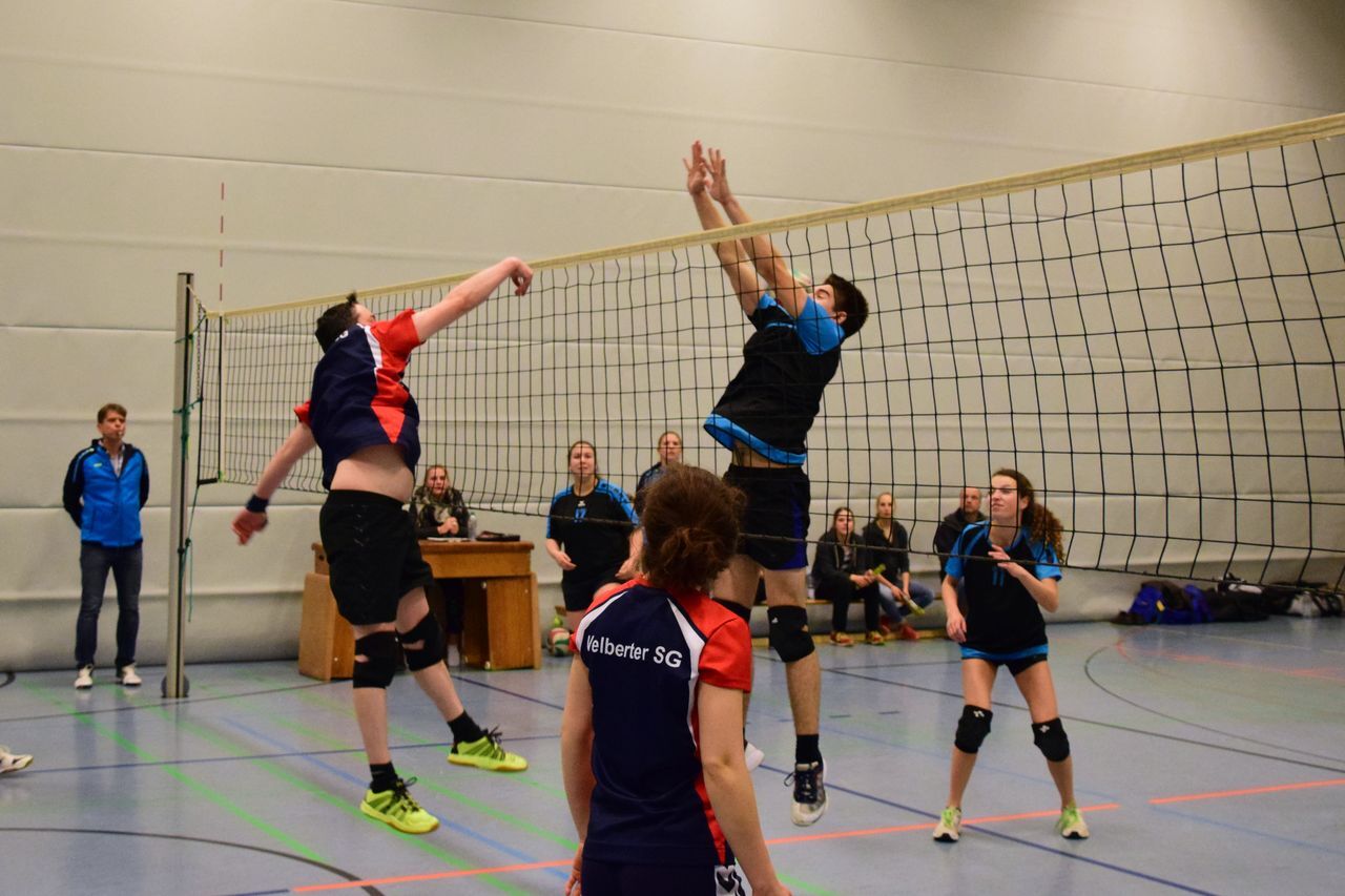 Zwei Volleyballspieler springen an einem Netz, während Zuschauer im Hintergrund auf einer Sporthalle sitzen.