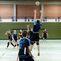 Volleyballspiel: Spieler springt zum Blocken, während andere konzentriert zuschauen. Netz und Sporthalle im Hintergrund.