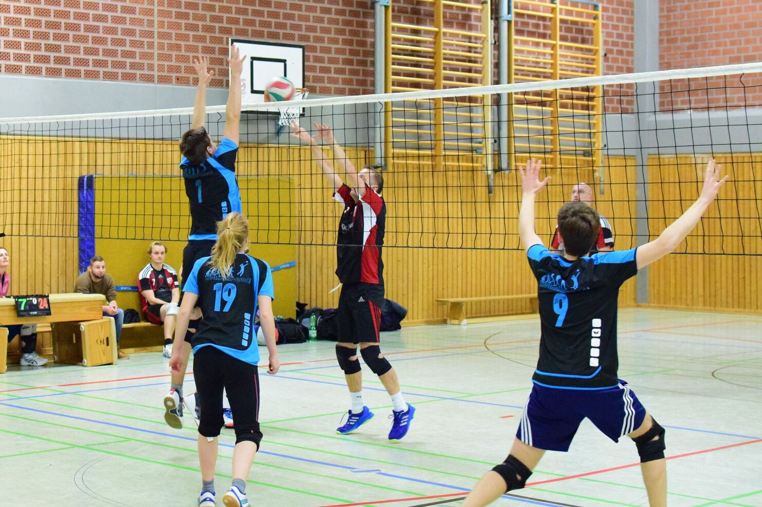 Volleyballspiel mit Spielern in blauen und roten Trikots, während ein Ball über das Netz geschlagen wird.