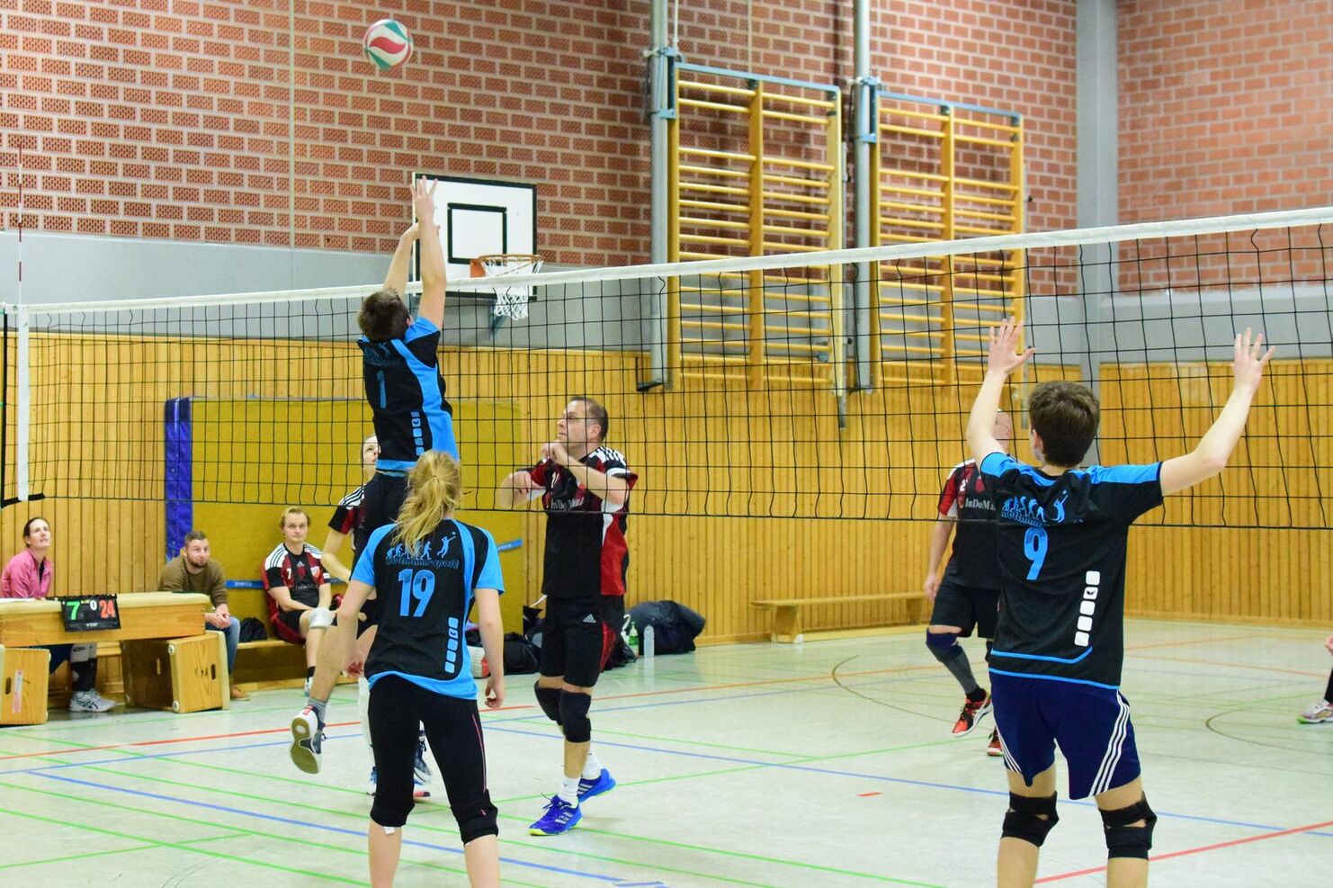 Volleyballspiel in einer Halle: Spieler mit blauem Trikot springt, um den Ball über das Netz zu blocken. Zuschauer im Hintergrund.