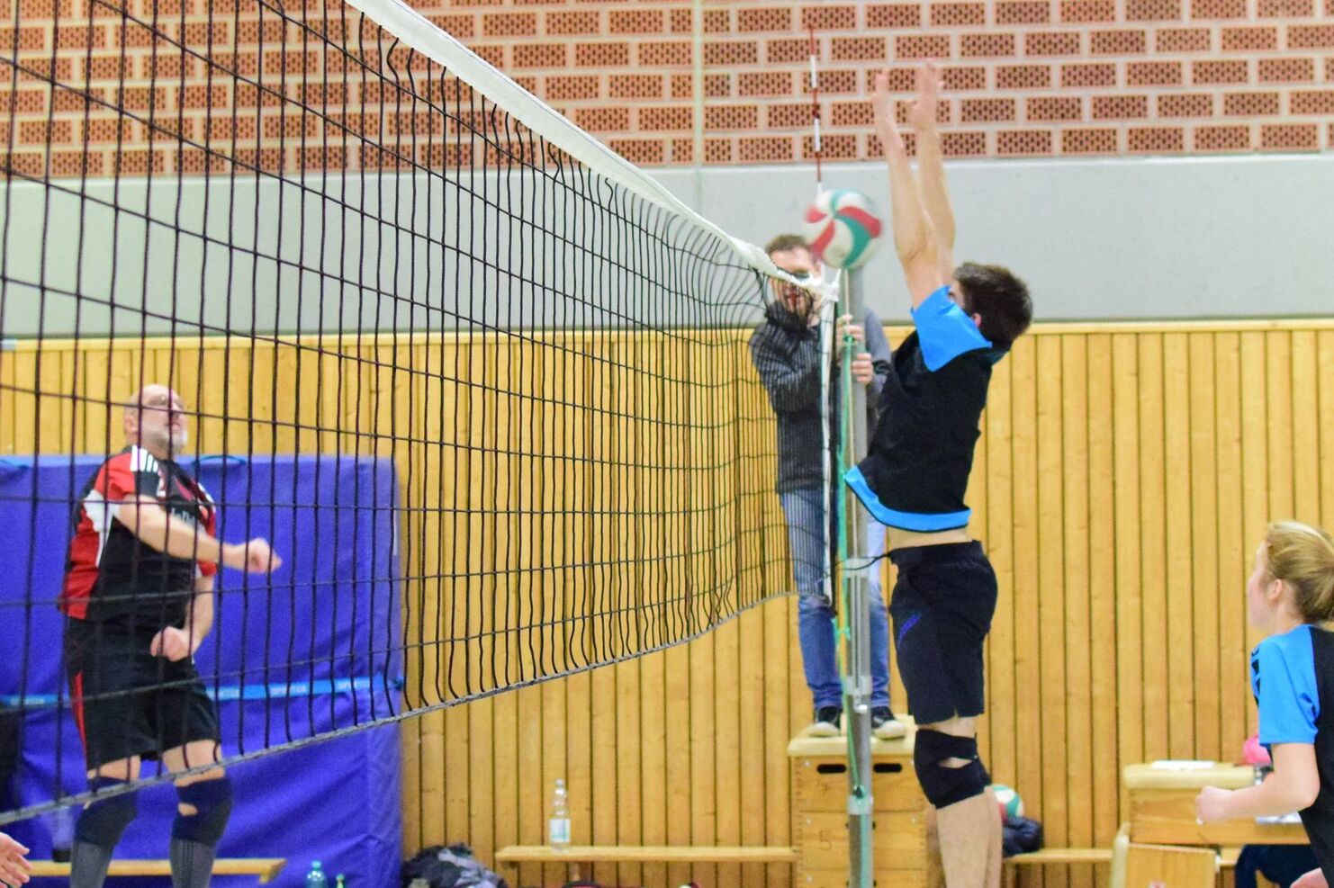 Zwei Volleyballspieler am Netz, einer springt, der andere bereitet sich auf den Angriff vor, Zuschauer im Hintergrund.