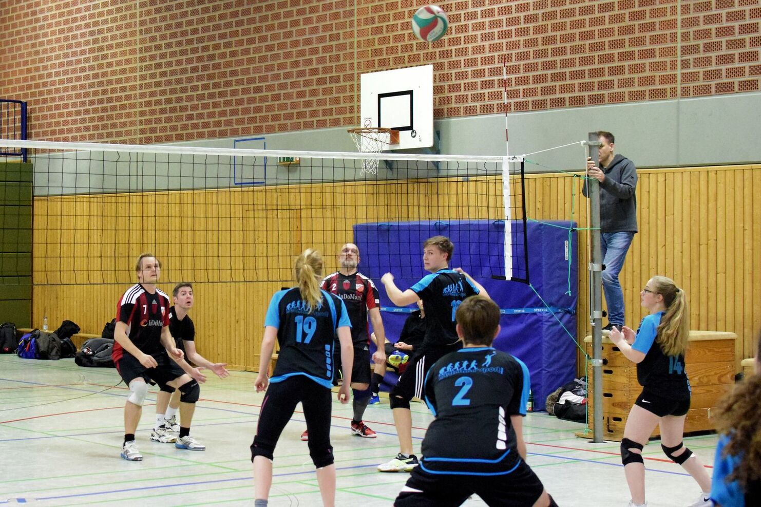 Volleyballspiel in einer Halle mit Spielern in schwarz-blauen Trikots und einem Ball, der über das Netz fliegt.