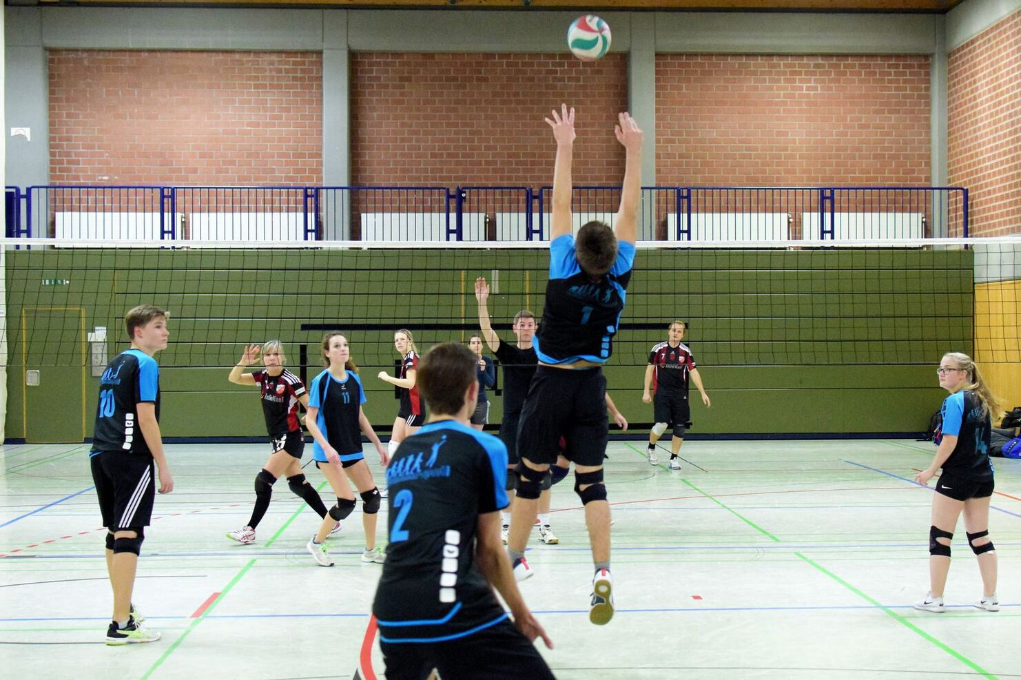 Volleyballspiel: Spieler springt zum Blocken, während andere konzentriert zuschauen. Netz und Sporthalle im Hintergrund.