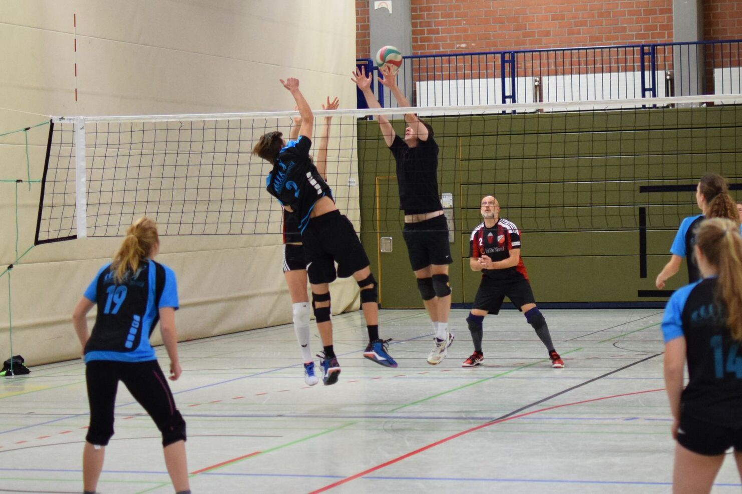 Zwei Spieler springen am Netz, um einen Volleyball während eines Spiels in einer Sporthalle zu blocken.