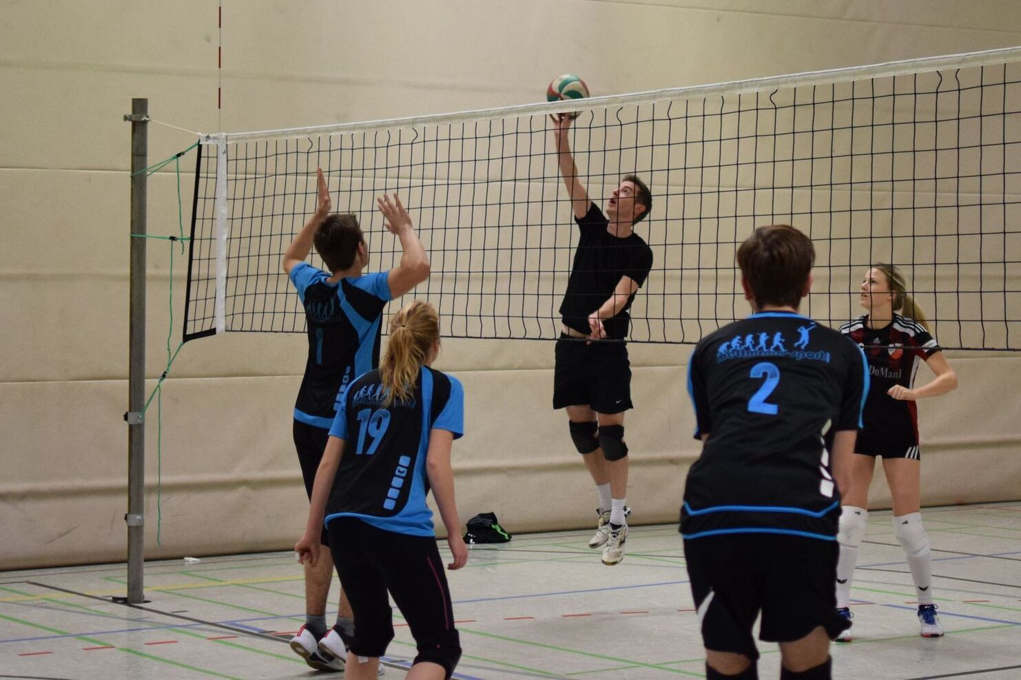 Spieler im schwarzen Trikot springt hoch, um den Volleyball über das Netz zu schlagen, während andere Spieler beobachten.
