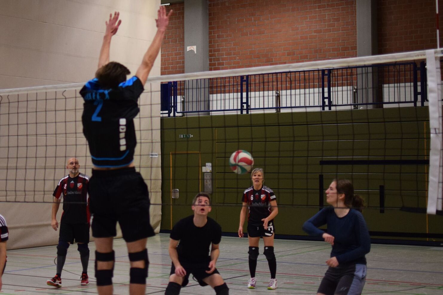 Volleyballspieler in Aktion, ein Spieler springt zum Block, während der Ball über das Netz fliegt. Zuschauer beobachten das Spiel.