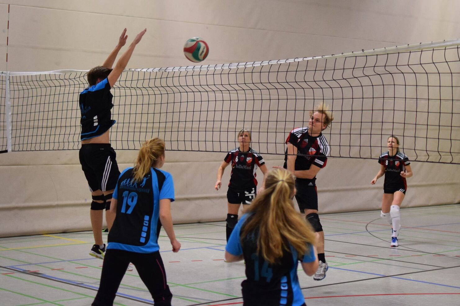 Mann springt zum Block, während ein Volleyball über das Netz fliegt; vier Spieler in blauen und schwarzen Trikots im Fokus.