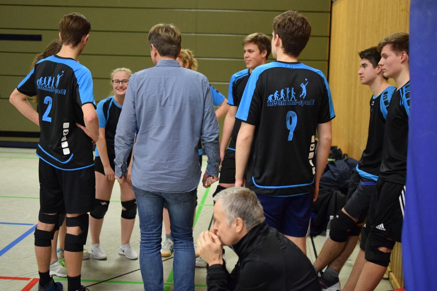 Gruppe von Volleyballspielern in Sportkleidung bespricht sich mit einem Trainer in einer Sporthalle.