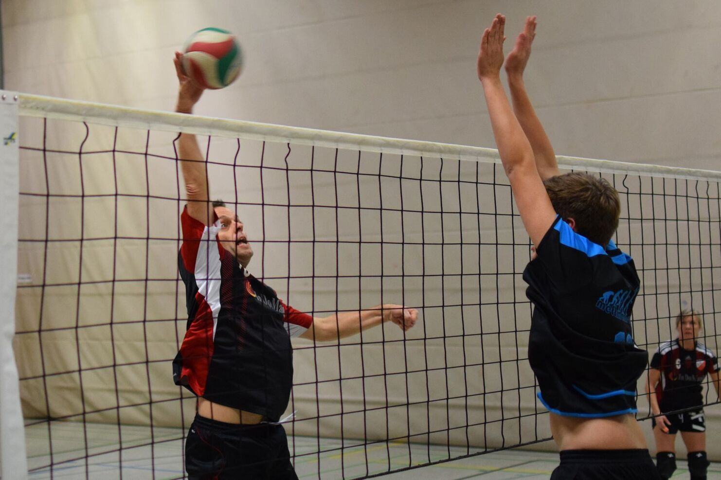 Ein Spieler springt zum Aufschlag, während ein Gegenspieler sich zum Blocken vorbereitet, beide in einer Halle.
