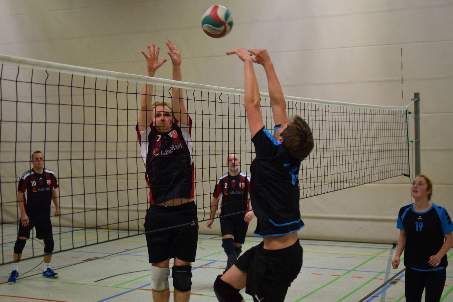 Zwei Volleyballspieler im Sprung am Netz, während ein Ball darüber fliegt; zwei weitere Spieler beobachten das Spiel.