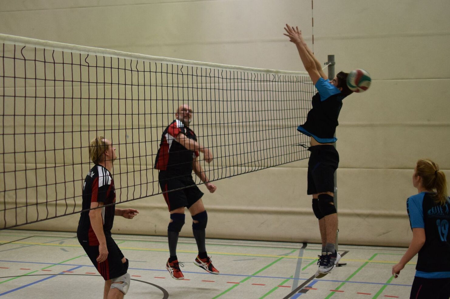 Viereckiger Volleyballplatz mit Spielern, die einen Ball am Netz ankämpfen. Zwei Spieler springen, ein weiterer beobachtet.