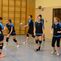 Sechs Volleyballspielende in Sportkleidung auf einem Hallenboden, bereit für das Training. Ein Spieler wirft den Ball.