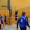 Spielszene in der Halle: Volleyballteam springt über das Netz, um den Ball zu schlagen. Mitglieder tragen blaue Trikots.