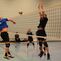 Ein Spieler springt, um einen Volleyball über das Netz zu schlagen, während andere Spieler in der Sporthalle beobachten.