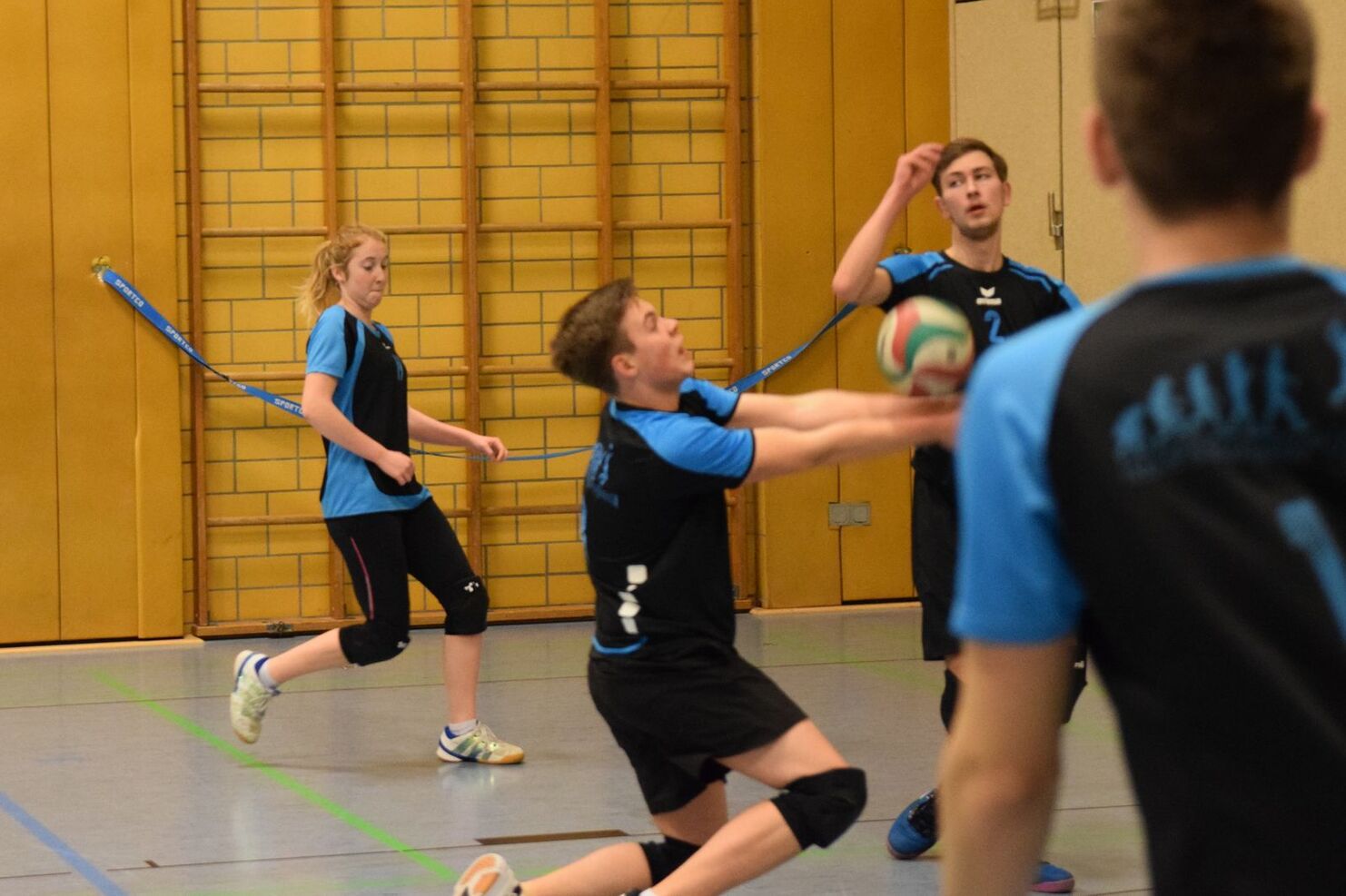 Vier Jugendliche spielen Volleyball in einer Sporthalle, während einer den Ball annehmen möchte.