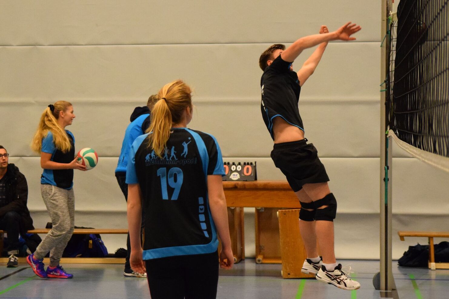 Junge Athleten in Sportkleidung spielen Volleyball in einer Turnhalle; einer springt zum Angriff.