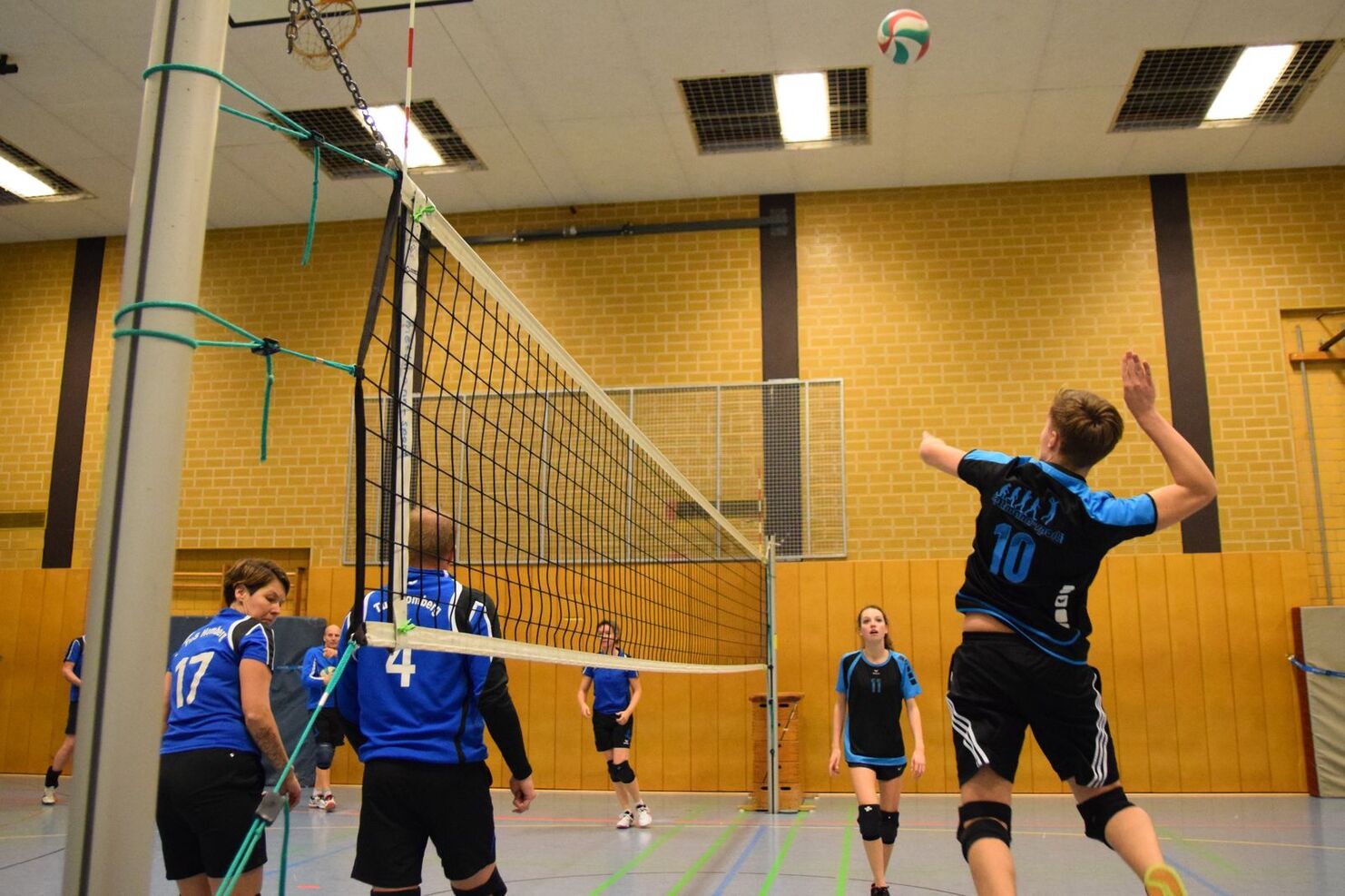Volleyballspieler im blauen Trikot springt zum Aufschlag. Teammitglieder beobachten auf einem Hallenboden.