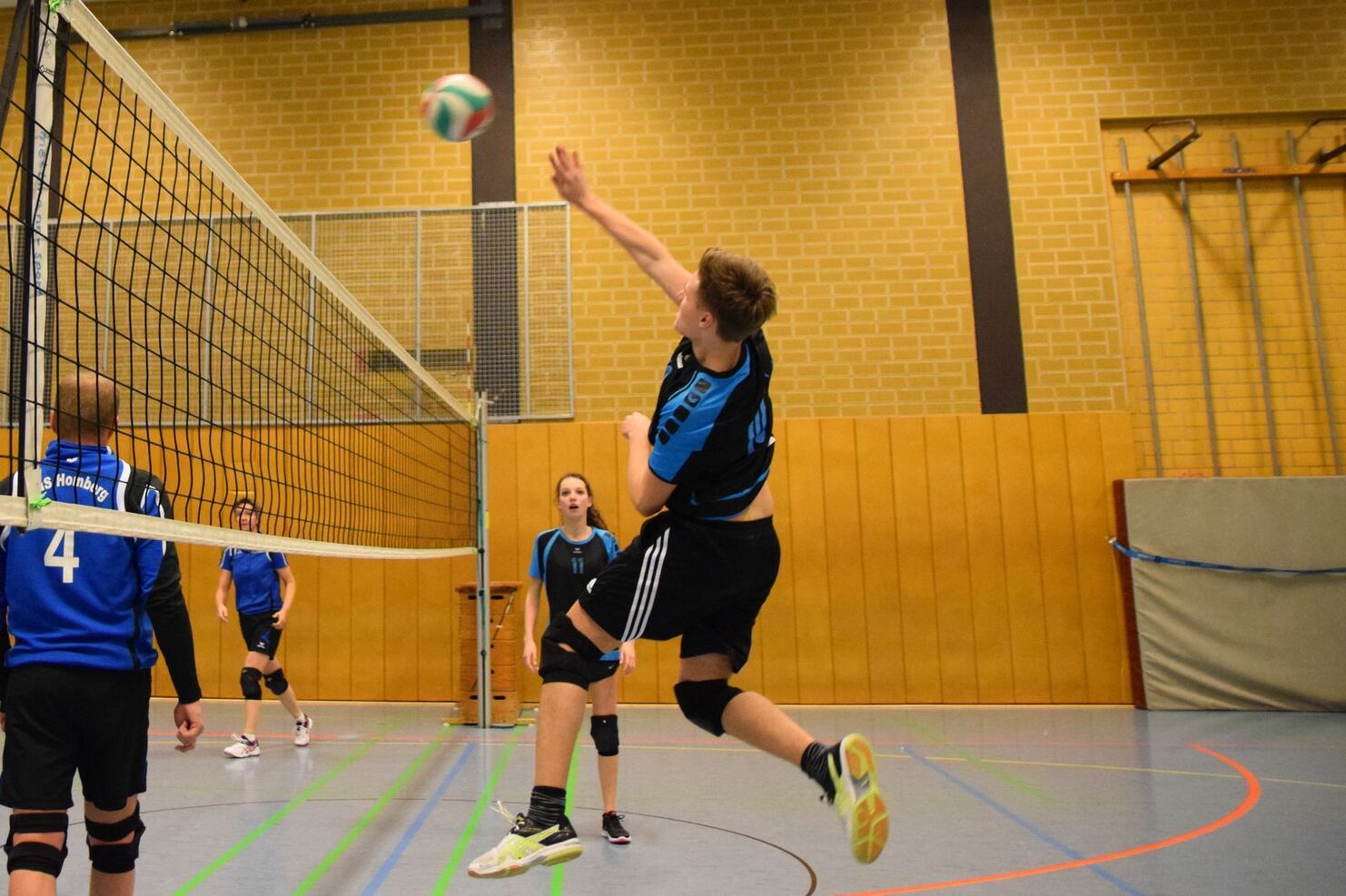 Spieler springt zum Aufschlag in einem Volleyballspiel, während andere Mannschaftskameraden aufmerksam zuschauen.