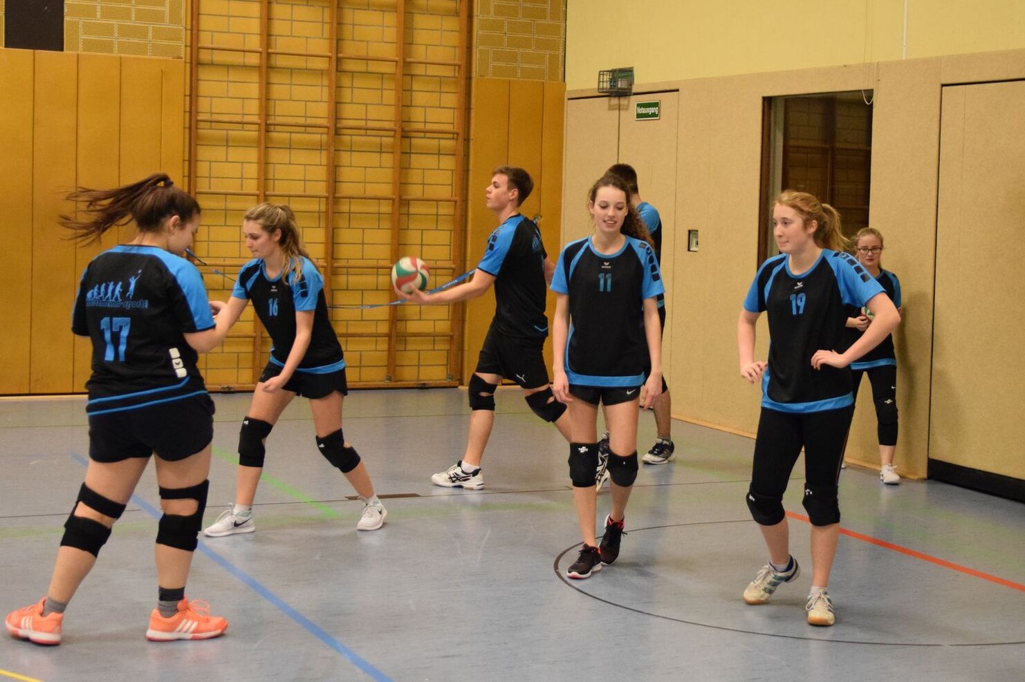 Sechs Volleyballspielende in Sportkleidung auf einem Hallenboden, bereit für das Training. Ein Spieler wirft den Ball.