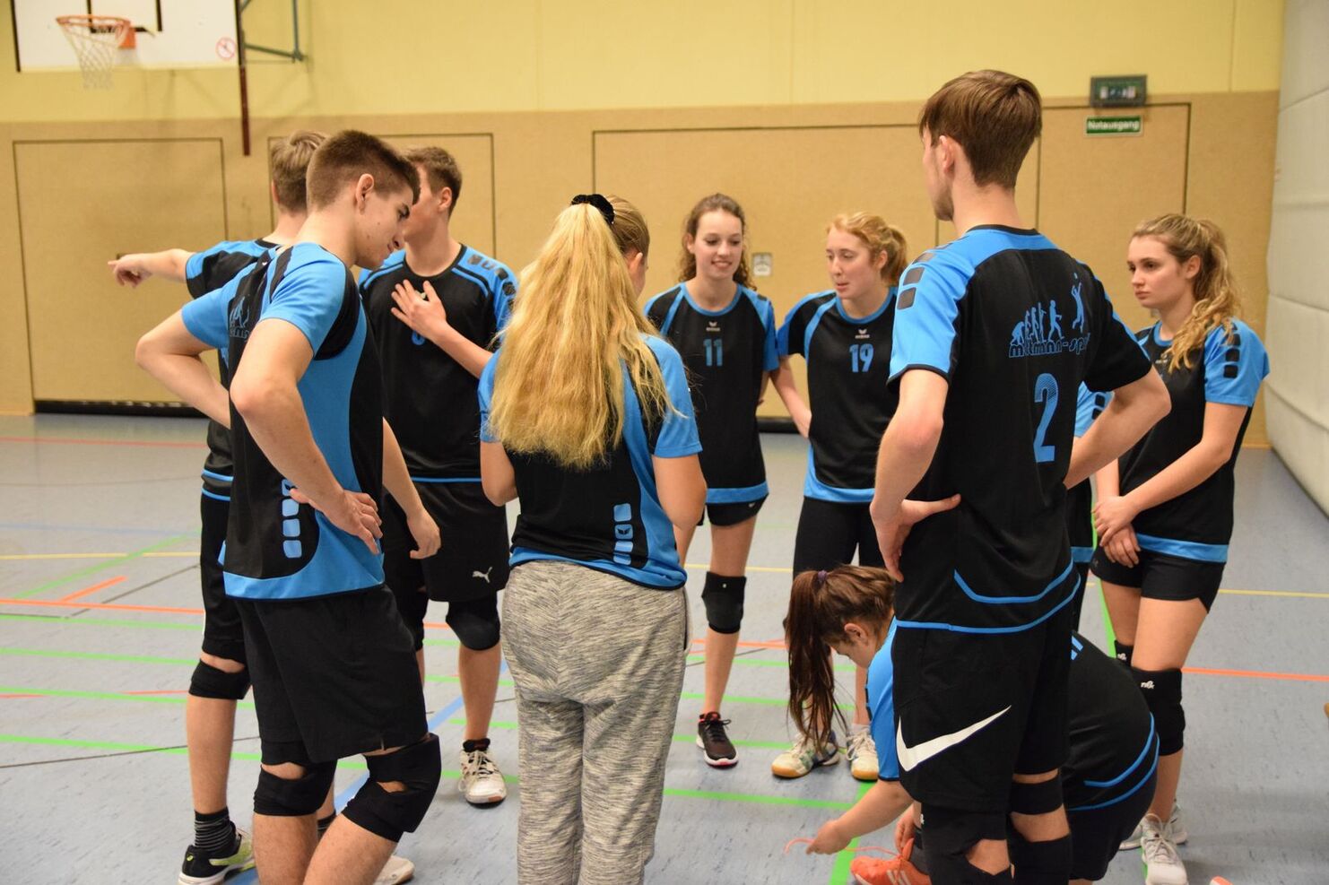Eine Gruppe von zehn Jugendlichen in Sportkleidung versammelt sich zur Besprechung in einer Sporthalle.
