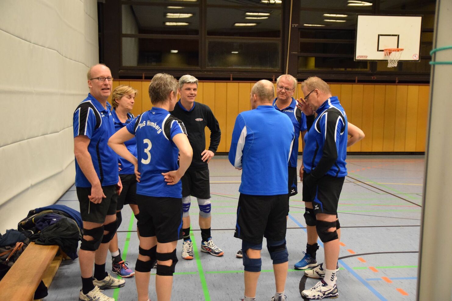 Gruppe von sieben Männern in blauen Sporttrikots versammelt sich in einer Sporthalle für eine Teambesprechung.