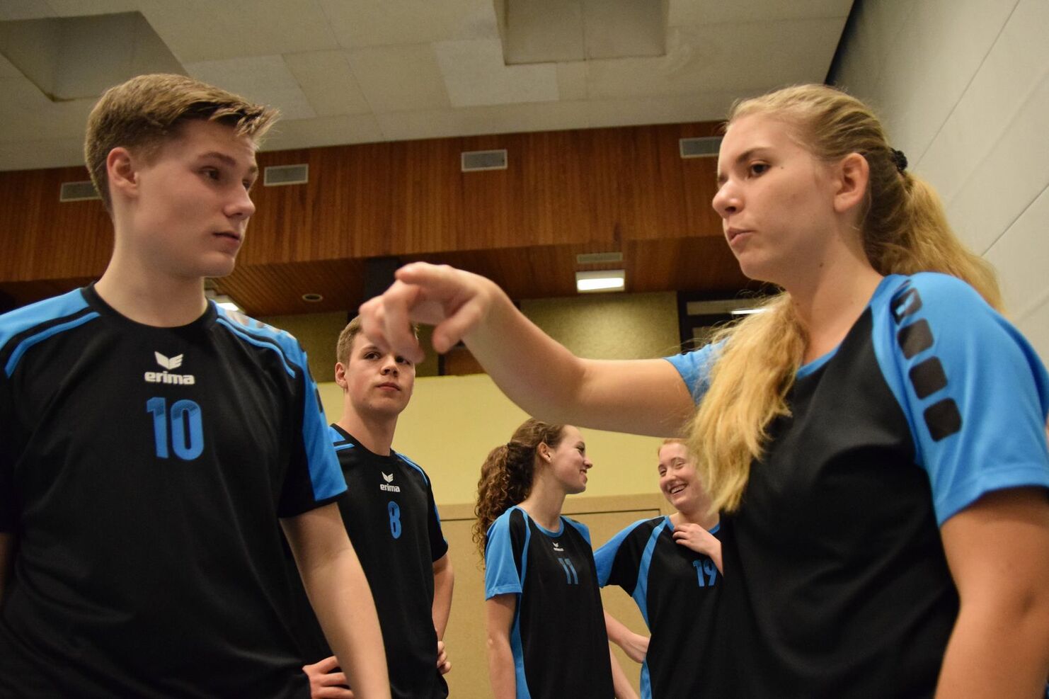 Vier Jugendliche in Sportbekleidung unterhalten sich in einer Halle, ein Junge hört aufmerksam zu, während eine Freundin spricht.