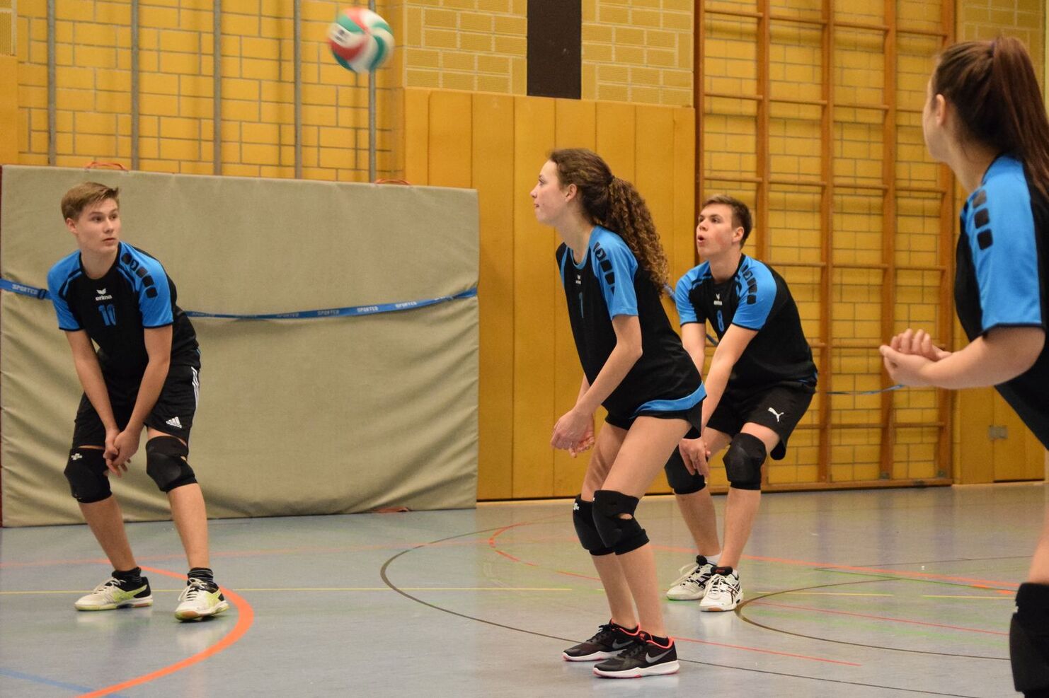 Vier Jugendliche in Sportkleidung stehen auf einem Volleyballfeld, während ein Ball in der Luft fliegt.