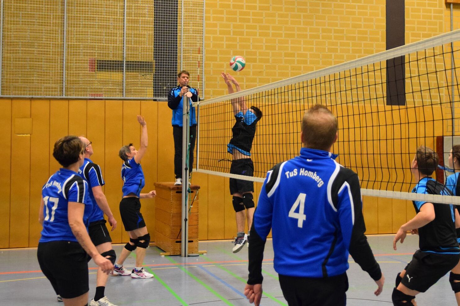 Spielszene in der Halle: Volleyballteam springt über das Netz, um den Ball zu schlagen. Mitglieder tragen blaue Trikots.