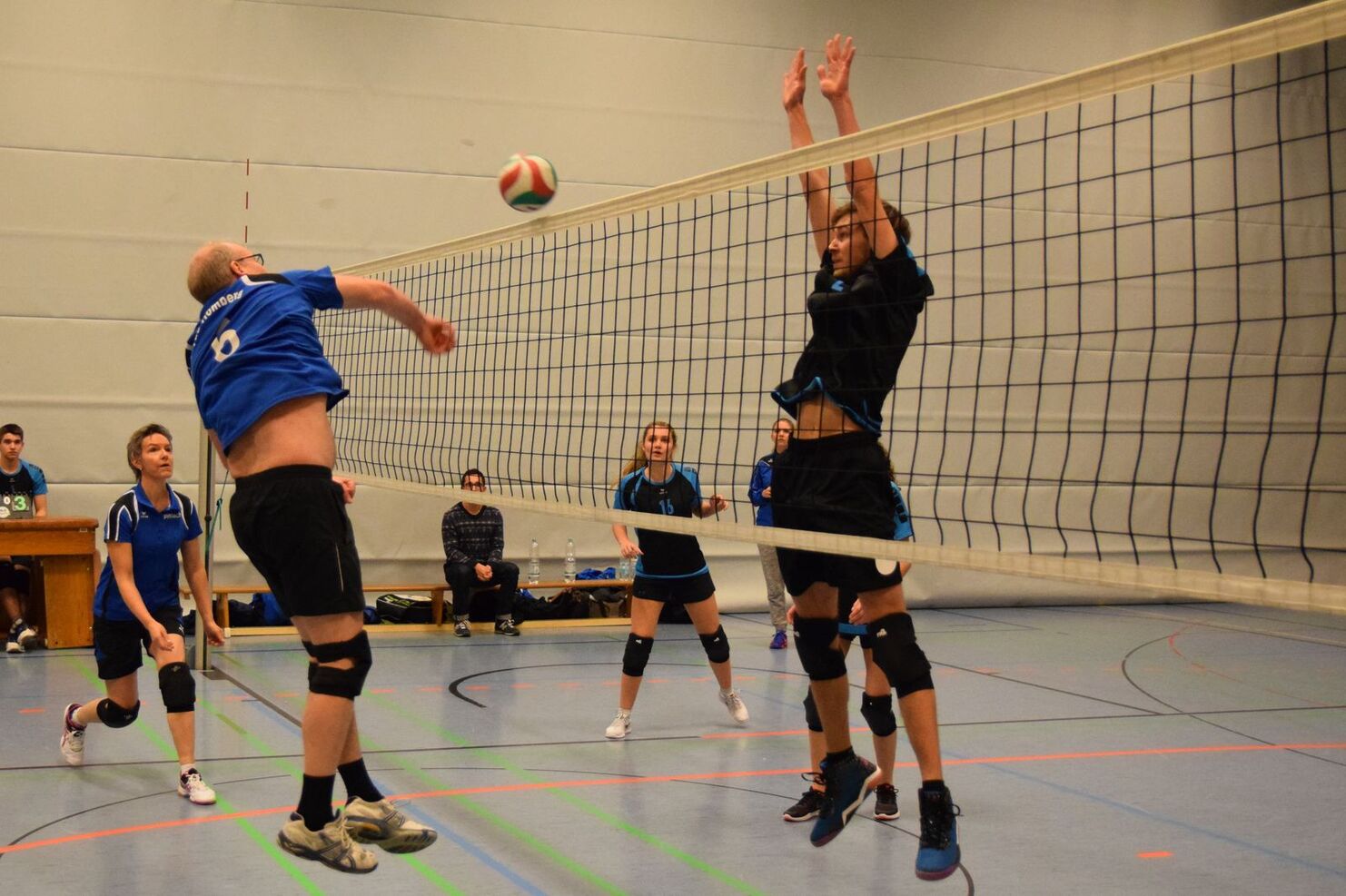 Ein Spieler springt, um einen Volleyball über das Netz zu schlagen, während andere Spieler in der Sporthalle beobachten.