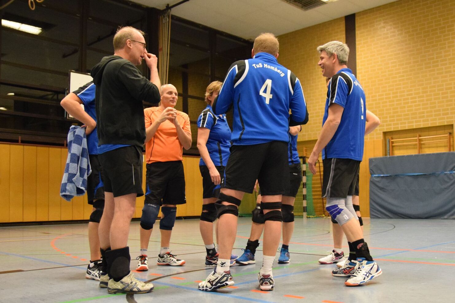 Sechs Sportler in blauen Trikots stehen um einen Spieler in orange, diskutieren in einer Sporthalle.