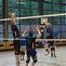 Zwei Volleyballspieler springen hoch, um einen Ball über das Netz zu schlagen, während andere Spieler im Hintergrund warten.
