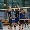 Zwei Spieler springen, um einen Volleyball über das Netz zu blocken, während weitere Spieler im Hintergrund agieren.