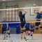 Zwei Volleyballspieler springen am Netz; im Hintergrund weitere Spieler in einer Sporthalle.