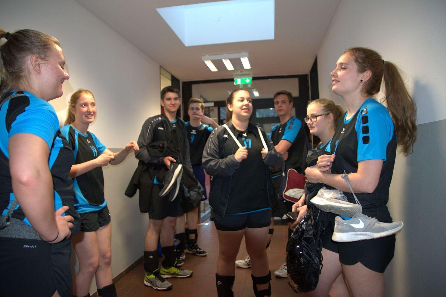 Gruppe junger Sportler in schwarzer und blauer Kleidung unterhält sich im Flur, einige halten Sportschuhe und Jacken.