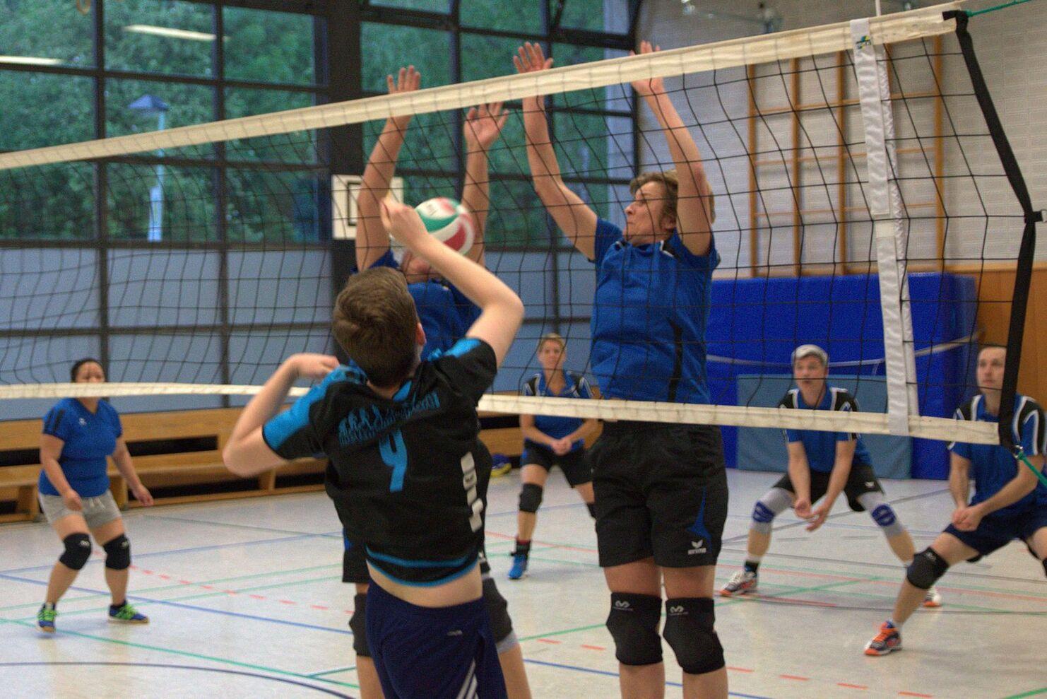 Volleyballspiel in einer Halle mit einem Spieler, der einen Aufschlag ausführt, während zwei Gegner am Netz blocken.