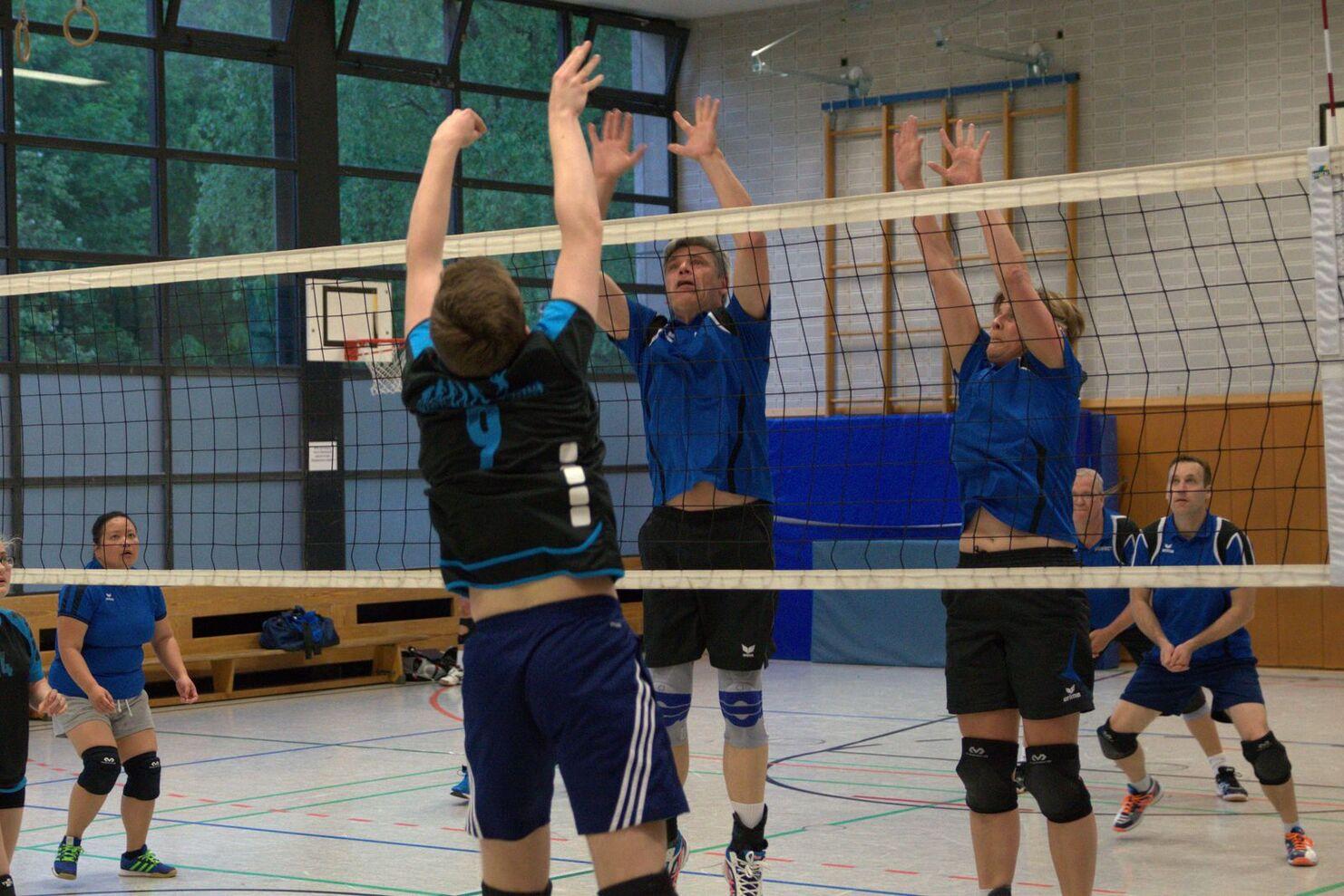 Zwei Spielerinnen blockieren einen Angreifer beim Volleyballspiel in einer Sporthalle. Zuschauer im Hintergrund.