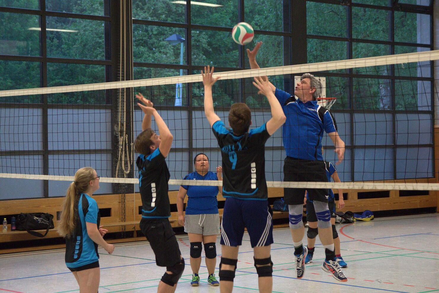 Spielszene in einer Volleyballhalle: Spieler springen hoch, um einen Ball über das Netz zu schlagen, während andere beobachten.