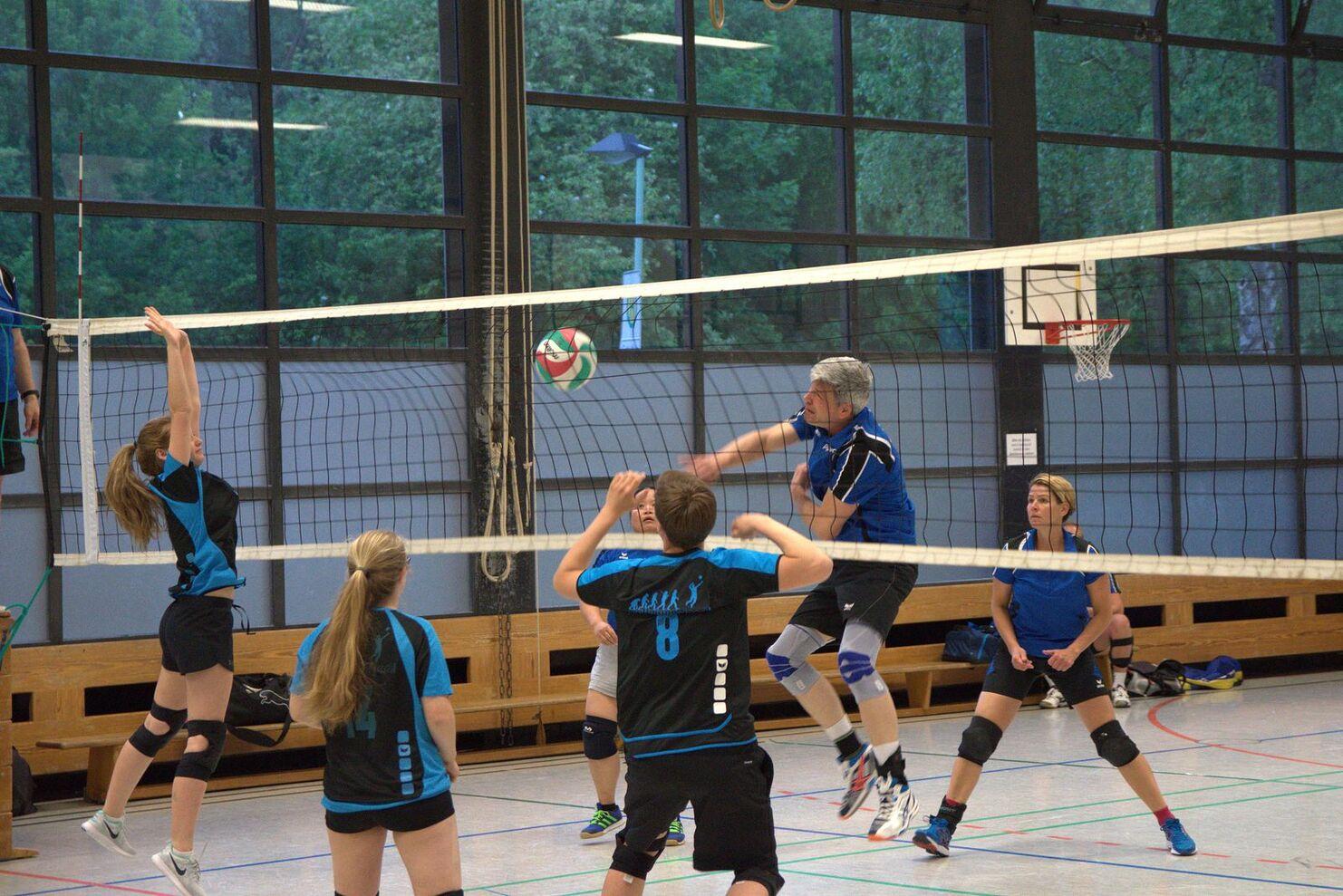 Volleyballspiel in einer Halle mit mehreren Spieler*innen, ein Spieler springt zum Angriff auf den Ball am Netz.