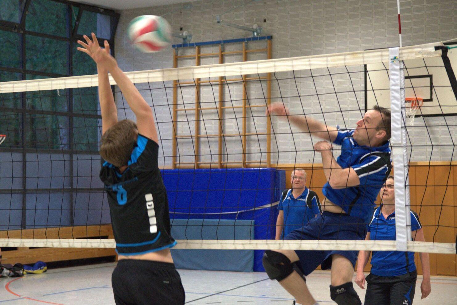Zwei Volleyballspieler im Wettkampf, einer springt zum Angriff, während der andere blockt. Zuschauer im Hintergrund.