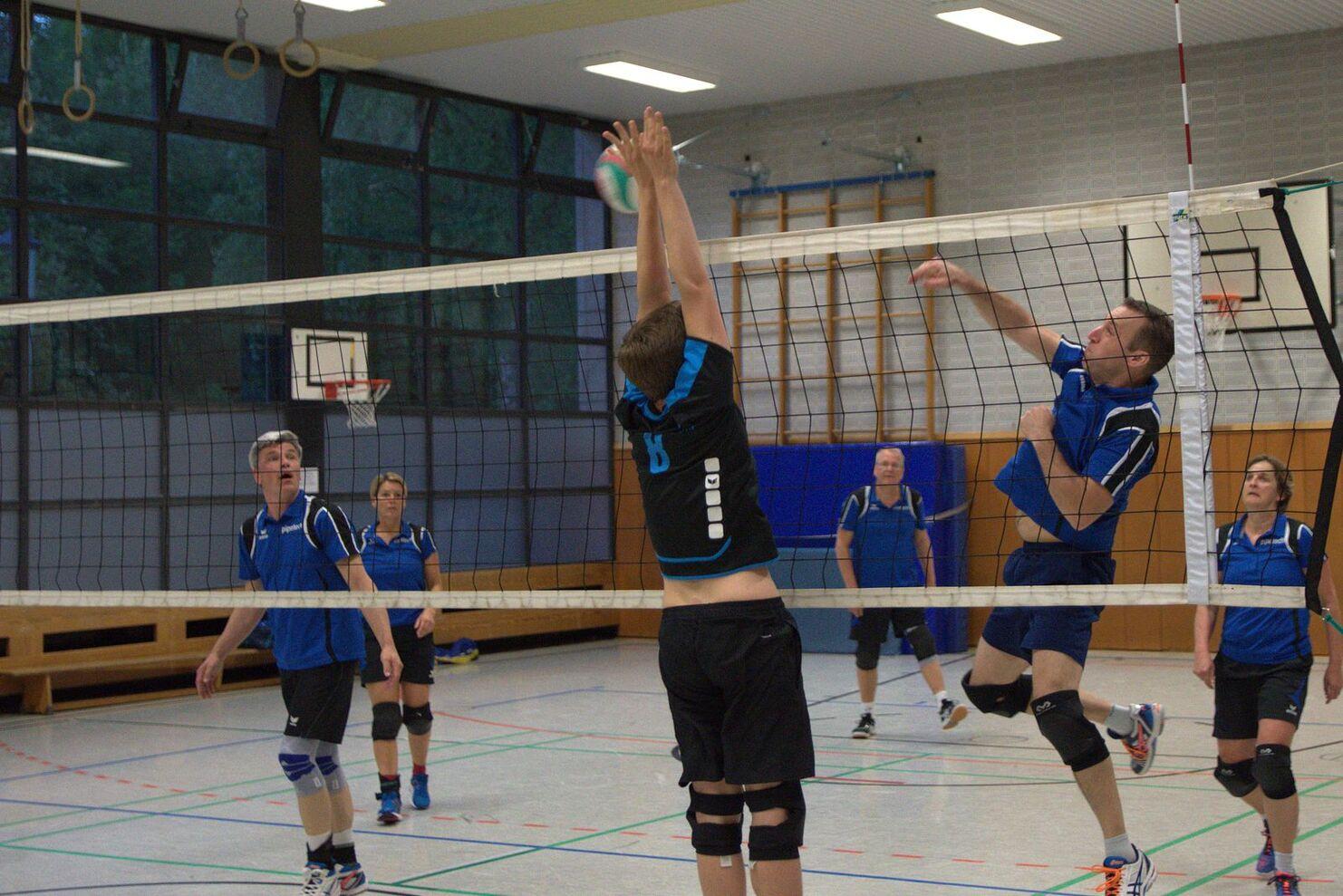 Volleyballspiel in einer Halle mit Spielern in blauen Trikots, einer springt, um den Ball über das Netz zu schlagen.