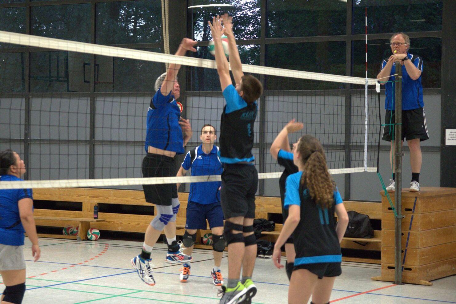 Volleyballspiel in einer Halle, ein Spieler springt zum Blocken, während andere Spieler im Hintergrund agieren.