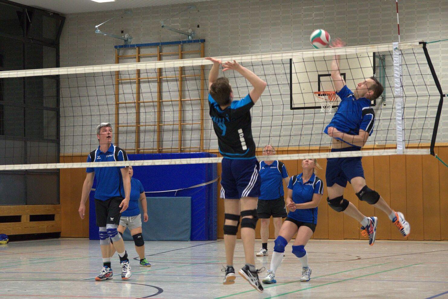 Zwei Volleyballspieler springen am Netz; im Hintergrund weitere Spieler in einer Sporthalle.
