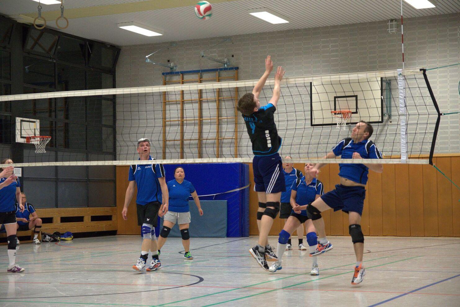 Volleyballspiel in einer Halle, ein Spieler springt, um den Ball über das Netz zu schlagen, während andere Zuschauer beobachten.