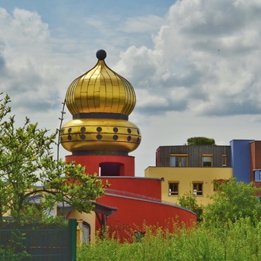 Bunte Gebäude mit einer goldenen Kuppel, umgeben von grünen Büschen und bewölktem Himmel.