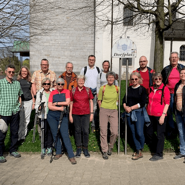 Gruppe von 16 Wanderern steht vor einem Hinweisschild "Dorfplatz" an einer Kirche im Freien.