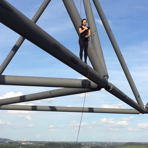 Frau steht auf einem großen Stahlträger mit Blick auf eine Landschaft und einen blauen Himmel. Seil ist sichtbar.